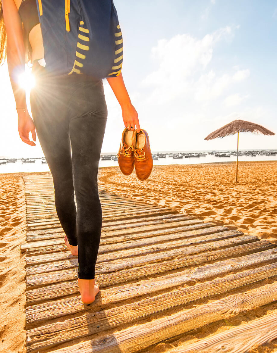 A woman in black tights, holding sneakers, walking on a boardwalk to the beach.